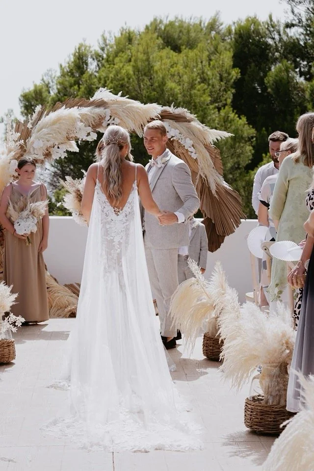 A couple exchanging vows during a wedding ceremony outdoors under a large, decorative arch made of dried pampas grass and palm leaves, with friends and family present.