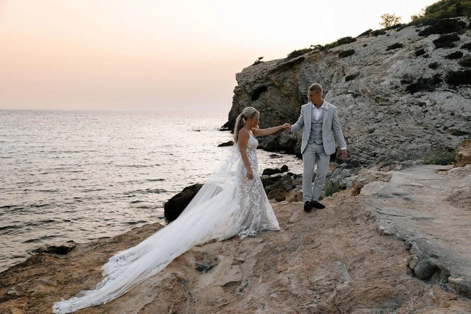 A bride and groom in wedding attire holding hands on rocky coast at sunset.