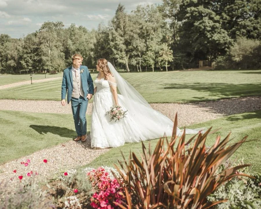 Bride and groom walking on a gravel path in a park, with trees and greenery in the background, during their wedding ceremony.