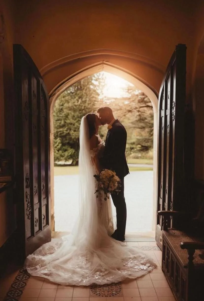 A bride and groom stand close together at the entrance of a church or chapel, holding a bouquet of flowers, with sunlight shining behind them and outdoor trees in the background.
