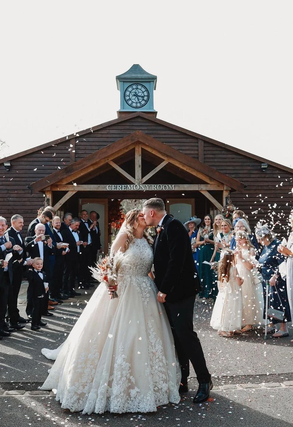 A newlywed couple kisses outside a wooden ceremony building, surrounded by wedding guests throwing confetti.