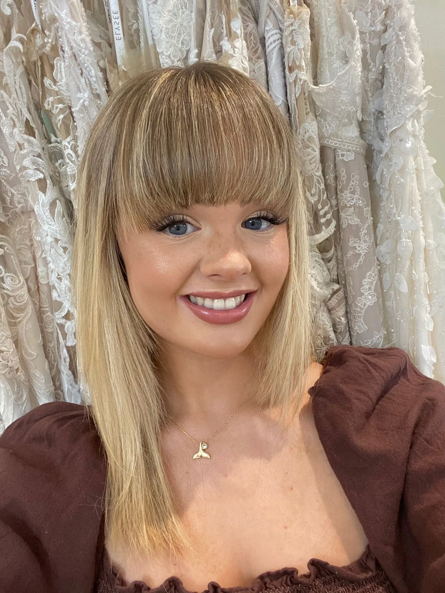 Young woman with blonde hair and bangs smiling in front of a background of lace and fabric. She is wearing a dark brown top with puffed shoulders and a gold necklace.