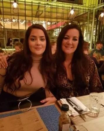 Two women sitting at a restaurant table, smiling, with plates, glasses, and silverware in front of them, inside a warmly lit dining area.