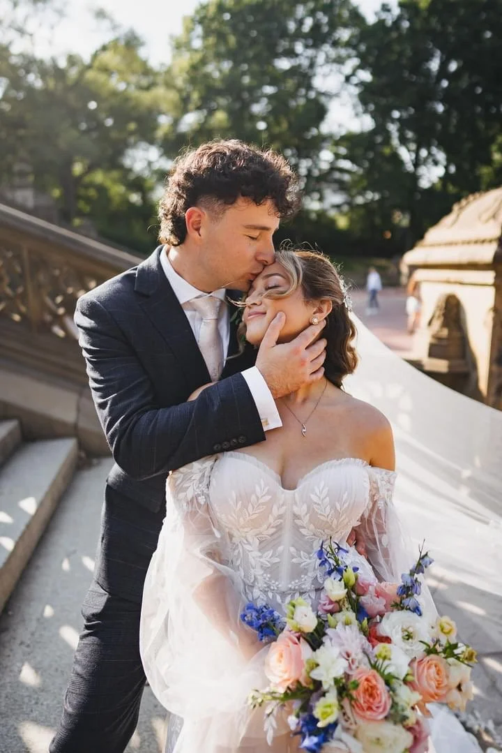 A groom in a dark suit gently kissing and holding the face of a bride in an off-the-shoulder white wedding dress, holding a colorful bouquet, outdoors on a staircase with trees in the background.