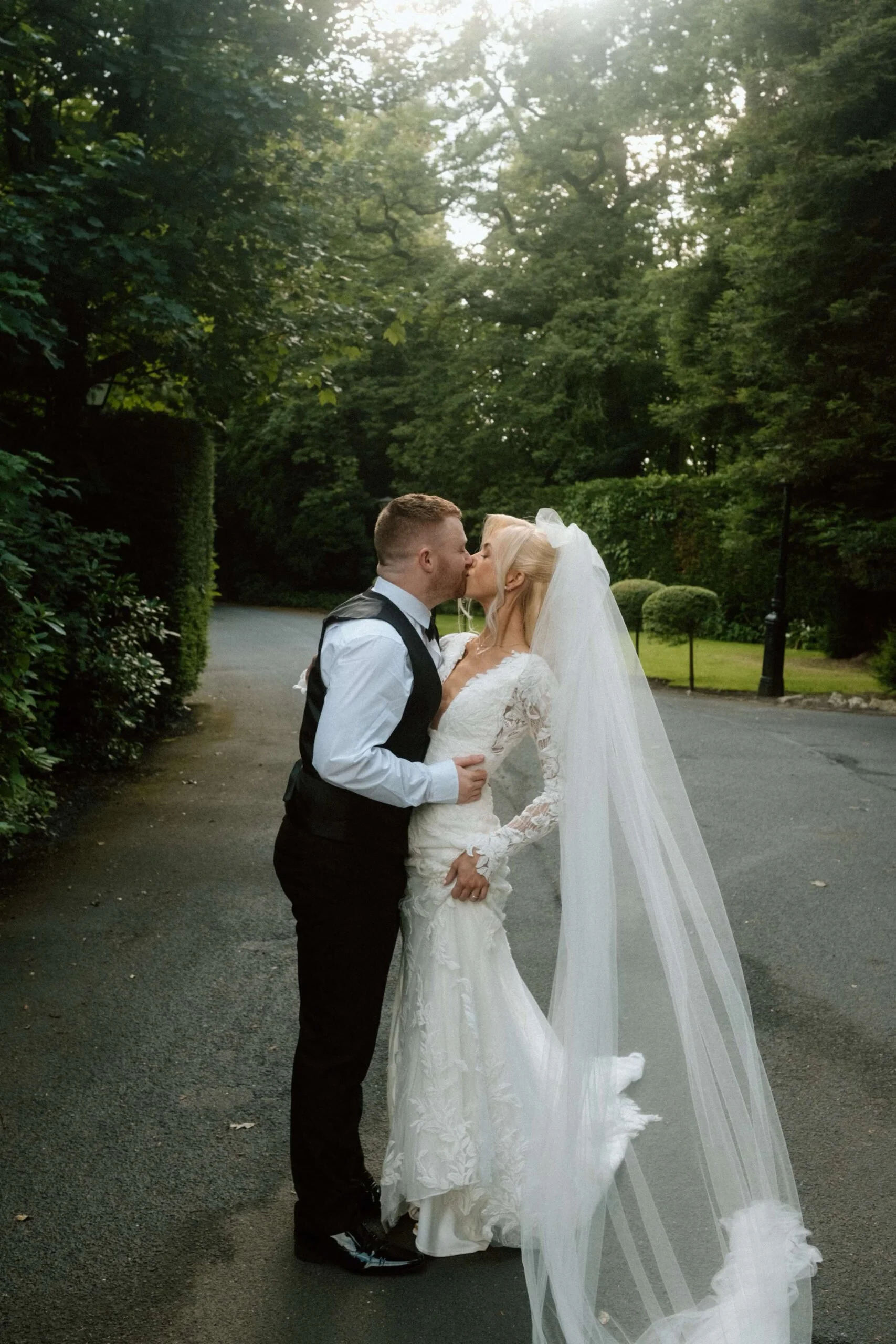 A bride and groom share a kiss outdoors, with trees and greenery in the background, on a paved path during a wedding wedding.