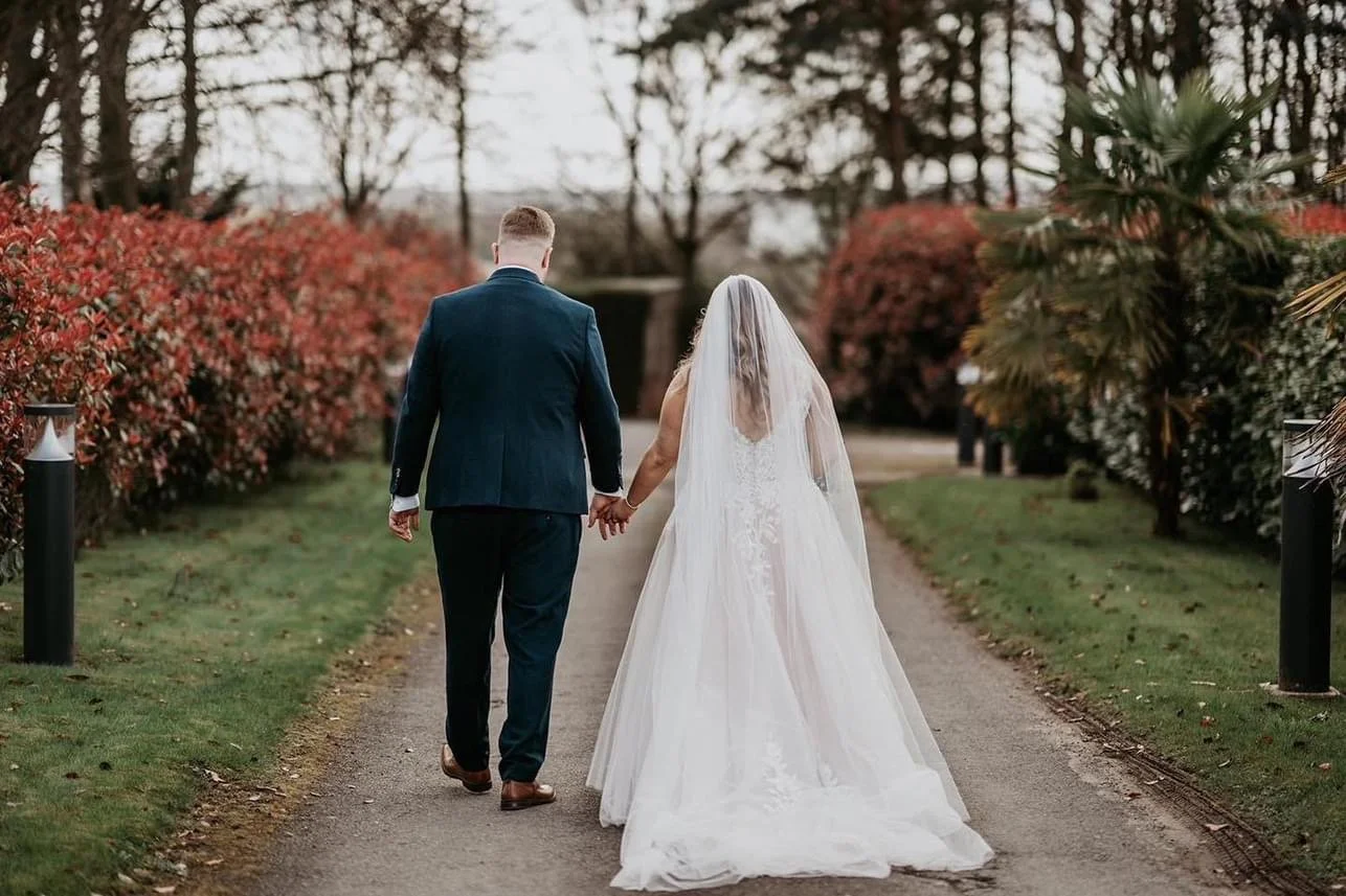 A bride and groom walking hand in hand down a garden path on their wedding day, surrounded by red and green foliage as they leave the venue.