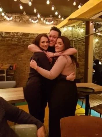 Three people embracing and smiling in a restaurant with warm lighting and string lights above.