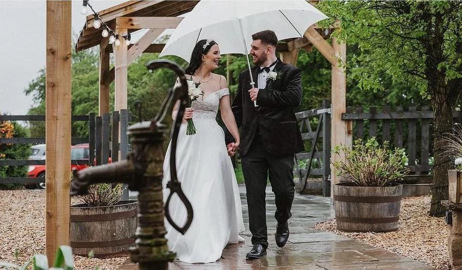 Bride and groom holding hands under an umbrella, smiling at each other outdoors during their wedding.