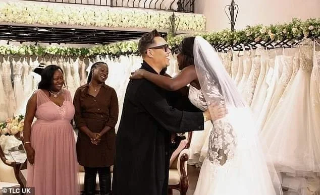 A bride and a man share a kiss during a wedding ceremony in a bridal shop, with two women standing and watching in the background.
