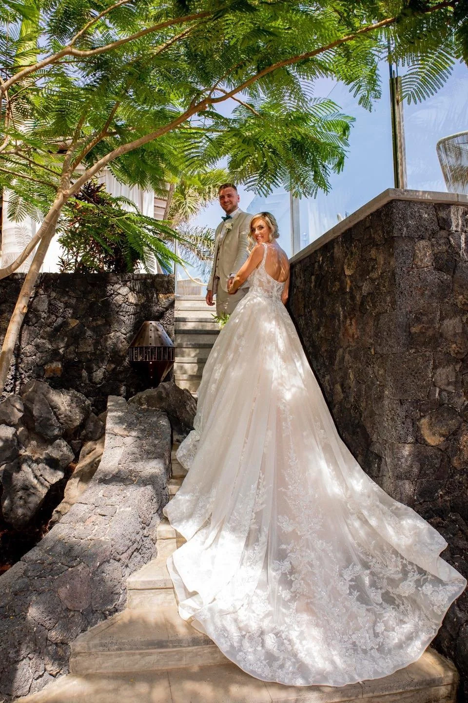 A bride and groom stand on stone steps outdoors, surrounded by greenery, with the bride in a white wedding dress and the groom in a light-colored suit. They are smiling and looking at the camera.