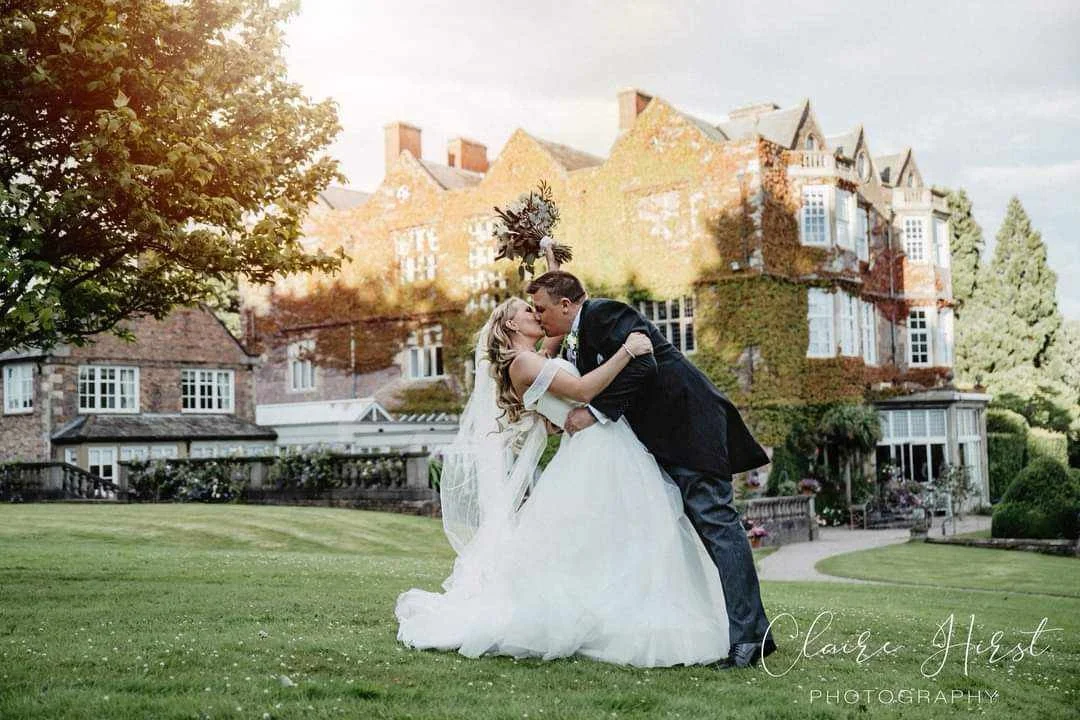 A bride and groom kiss on a lawn in front of a historic mansion with ivy-covered walls and multiple windows.