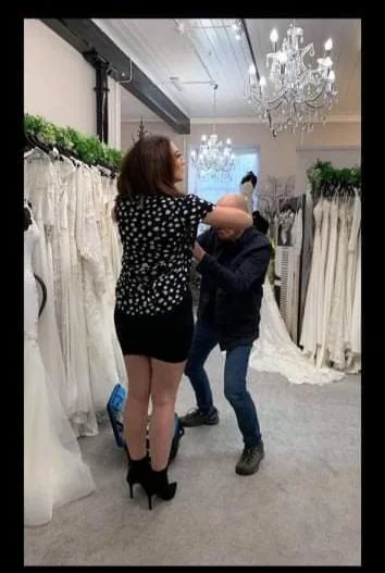 A woman in black high heels and a polka dot dress is playing with a small dog in a room decorated with chandeliers and green plants, with another person standing nearby.