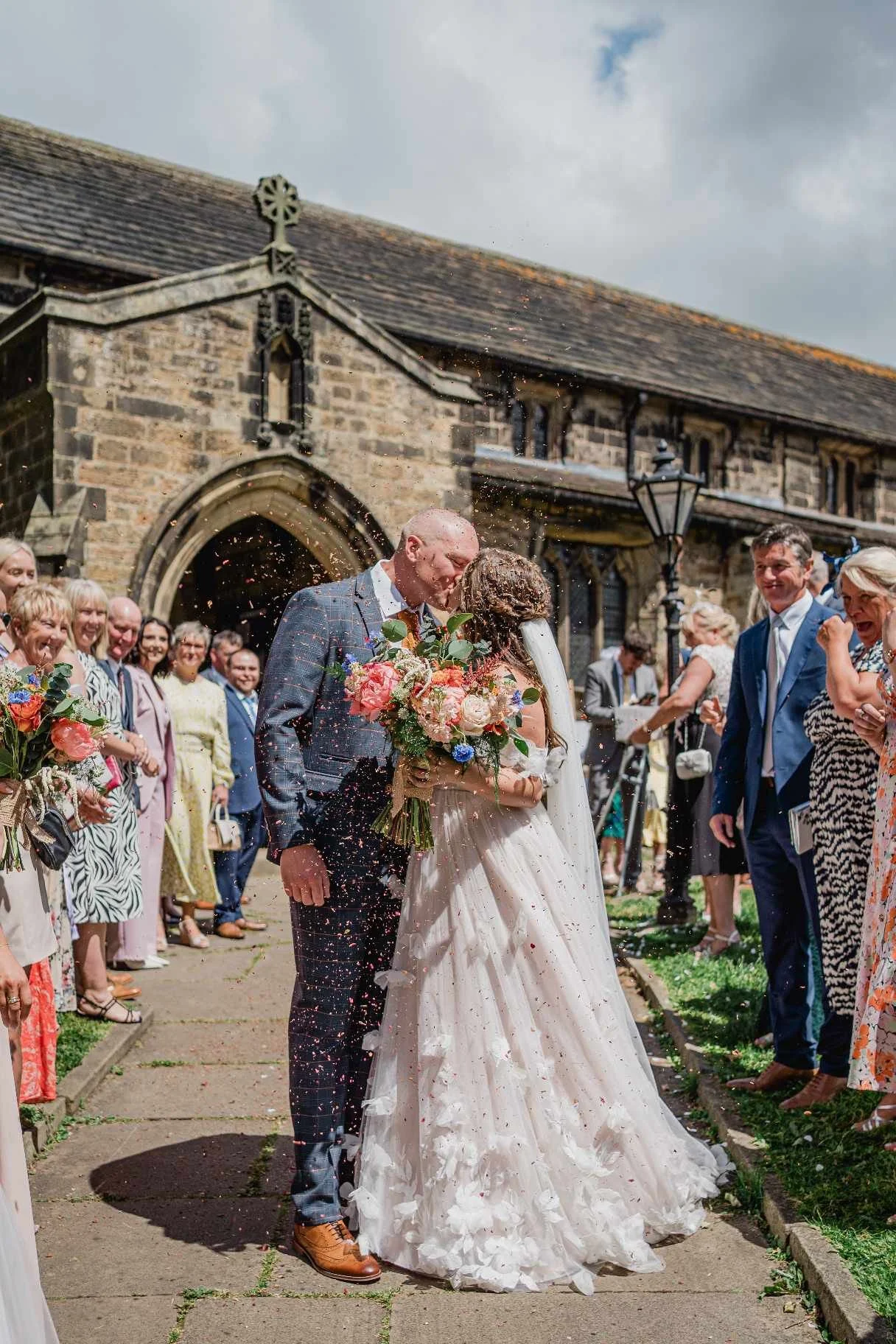 A couple kissing during their wedding ceremony outside a church, with guests celebrating around them.
