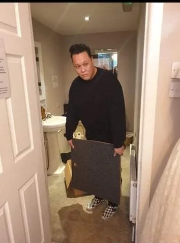 A man standing in a doorway holding a large package in a room with a bathroom in the background.