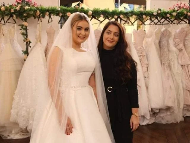 Two women, one in a wedding dress and the other in black, standing together in front of a rack of dresses.