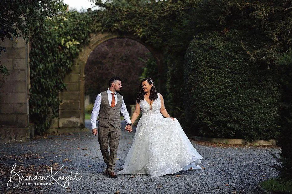 A bride and groom walking hand in hand outdoors, surrounded by greenery and an arched stone structure, during their wedding.