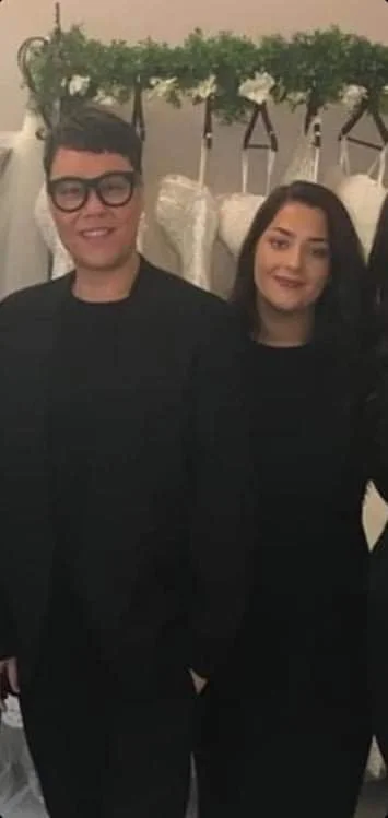 Two people standing indoors, smiling, with a decorative flower and greenery arrangement behind them.