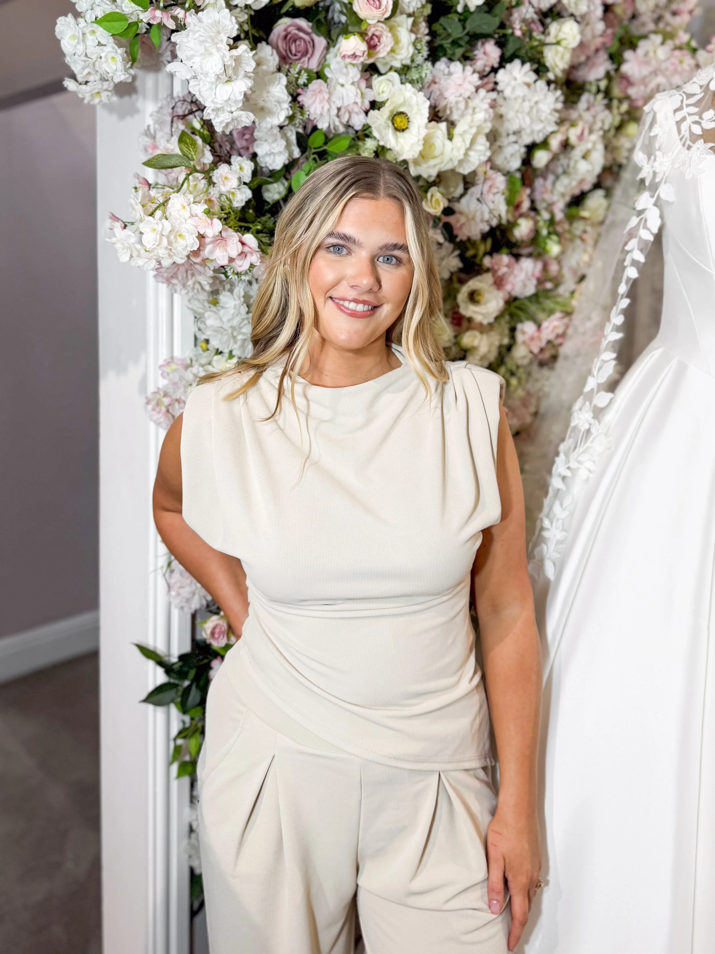 Close-up of a smiling woman with blonde hair and blue eyes in a bridal boutique, trying on a black dress with wedding dresses in the background.