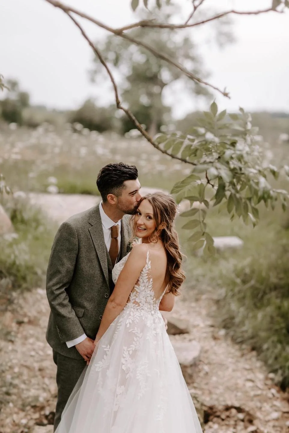 A bride and groom on their wedding day outdoors, with the groom kissing the bride on her forehead and the bride smiling.