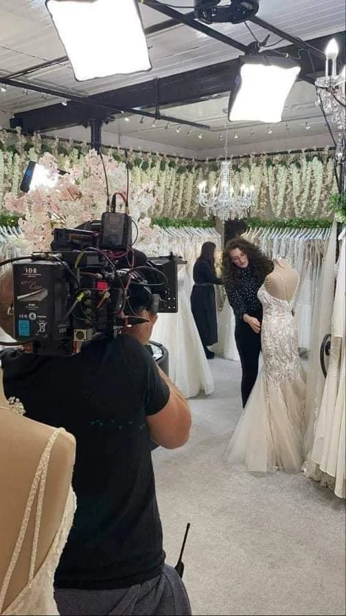 A woman with curly hair fitting a wedding dress on a mannequin in a bridal boutique, with a photographer filming her and another woman in the background, and floral decorations hanging from the ceiling.