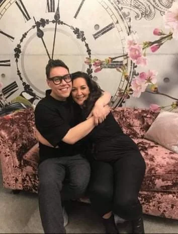 A man and woman are sitting on a pink velvet sofa, hugging and smiling. Behind them is a large wall clock surrounded by pink flowers and decorative elements.