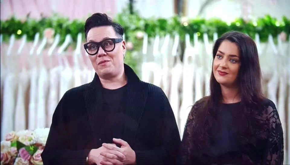 A man with black hair, glasses, and a black outfit stands beside a woman with long dark hair and a black lace dress, standing outdoors with floral decorations in the background.