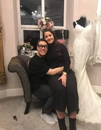 Two women sitting on a striped armchair indoors, with a window and a wedding dress on a mannequin in the background, and floral decorations above.