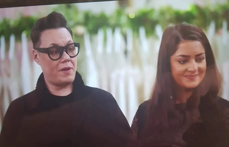 Two women standing outdoors with trees and white fencing in the background.