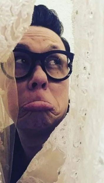 Person with glasses looking upward with a sad or disappointed expression, peeking through a lace curtain.
