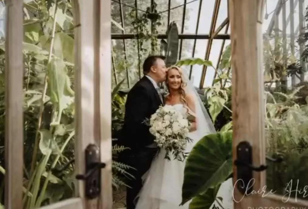A couple on their wedding day standing inside a greenhouse filled with large leafy plants. The groom is kissing the bride on the forehead, and the bride is smiling and holding a bouquet of white flowers. The greenhouse has glass walls and a wooden do