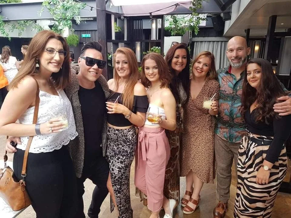 A group of nine friends at a social gathering, smiling and holding drinks, inside a decorated venue with plants and black furniture.