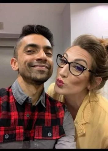 A man and a woman taking a selfie indoors, smiling and making a kiss face. The man has short dark hair, a beard, and is wearing a plaid shirt. The woman has glasses, styled hair, and is wearing a yellow jacket.