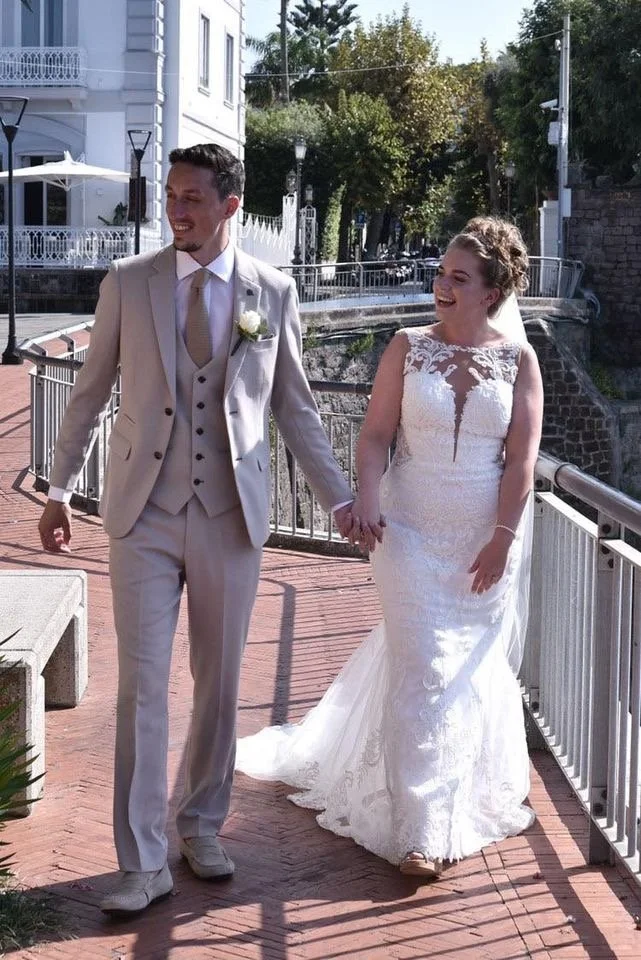 A newlywed couple holding hands and smiling, walking outdoors on a brick pathway in a sunny urban setting with green trees, buildings, and a bridge in the background.