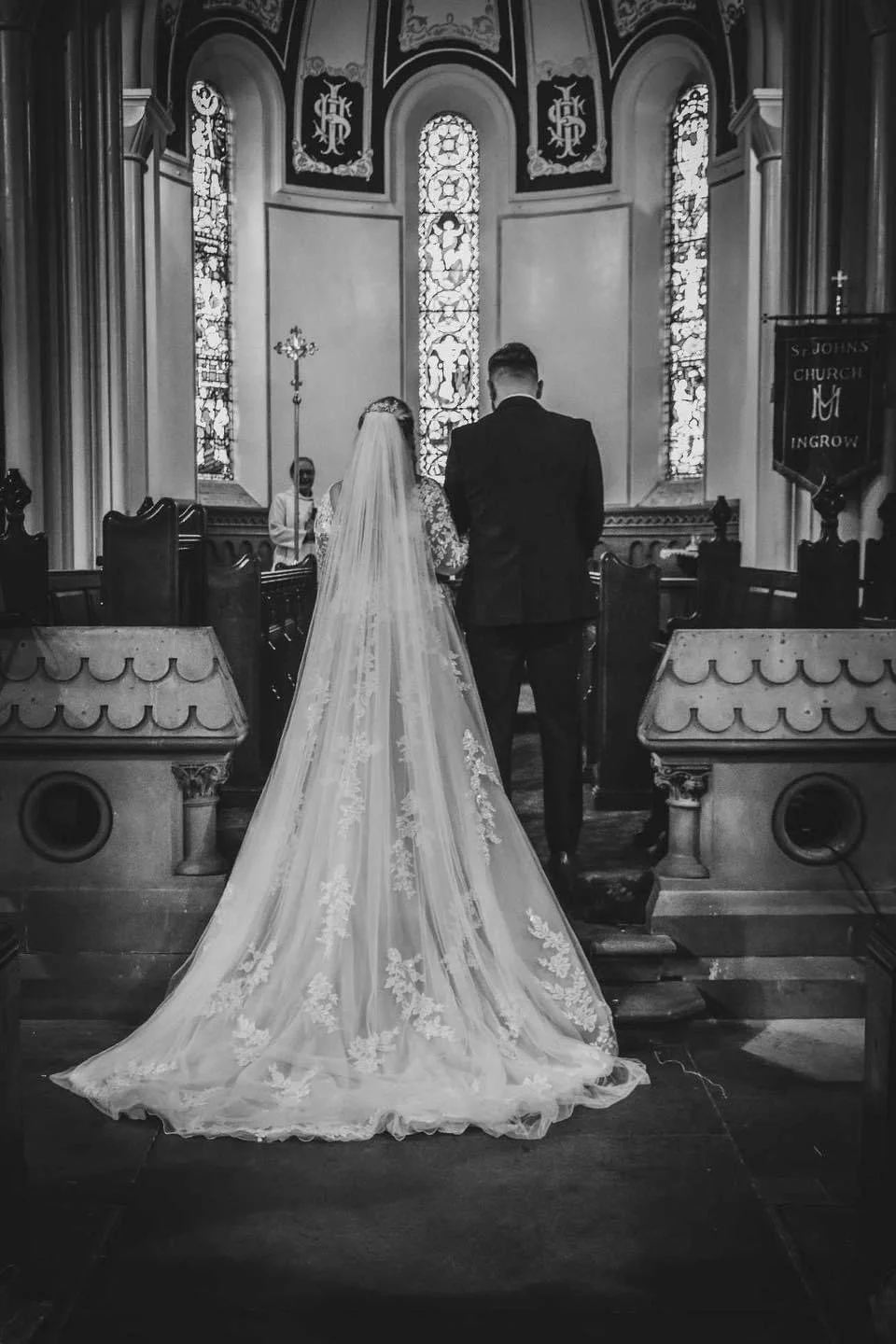A bride and groom standing at the altar in a church during their wedding ceremony, facing the priest, with stained glass windows behind them.