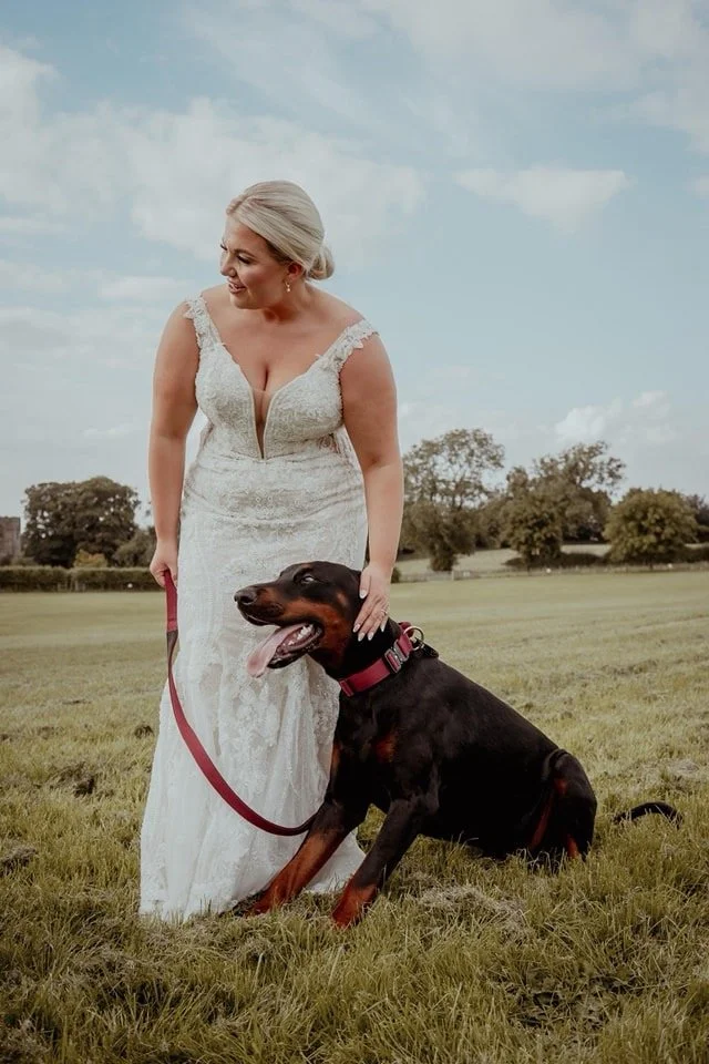 A woman in a white lace wedding dress outdoors with a large black and brown dog on a red leash in a grassy field with trees and cloudy sky in the background.