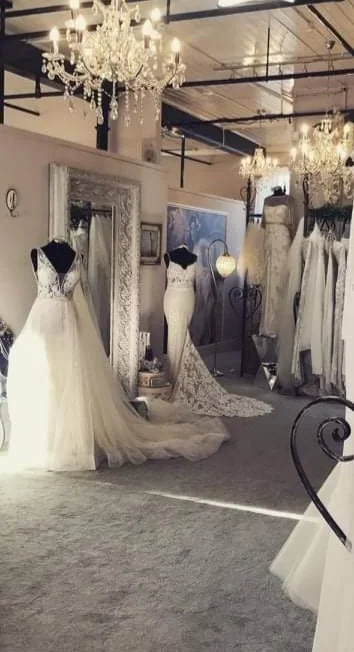 A bridal boutique with wedding dresses displayed on mannequins and hanging on racks, with chandeliers hanging from the ceiling, decorative mirrors, and elegant decor.