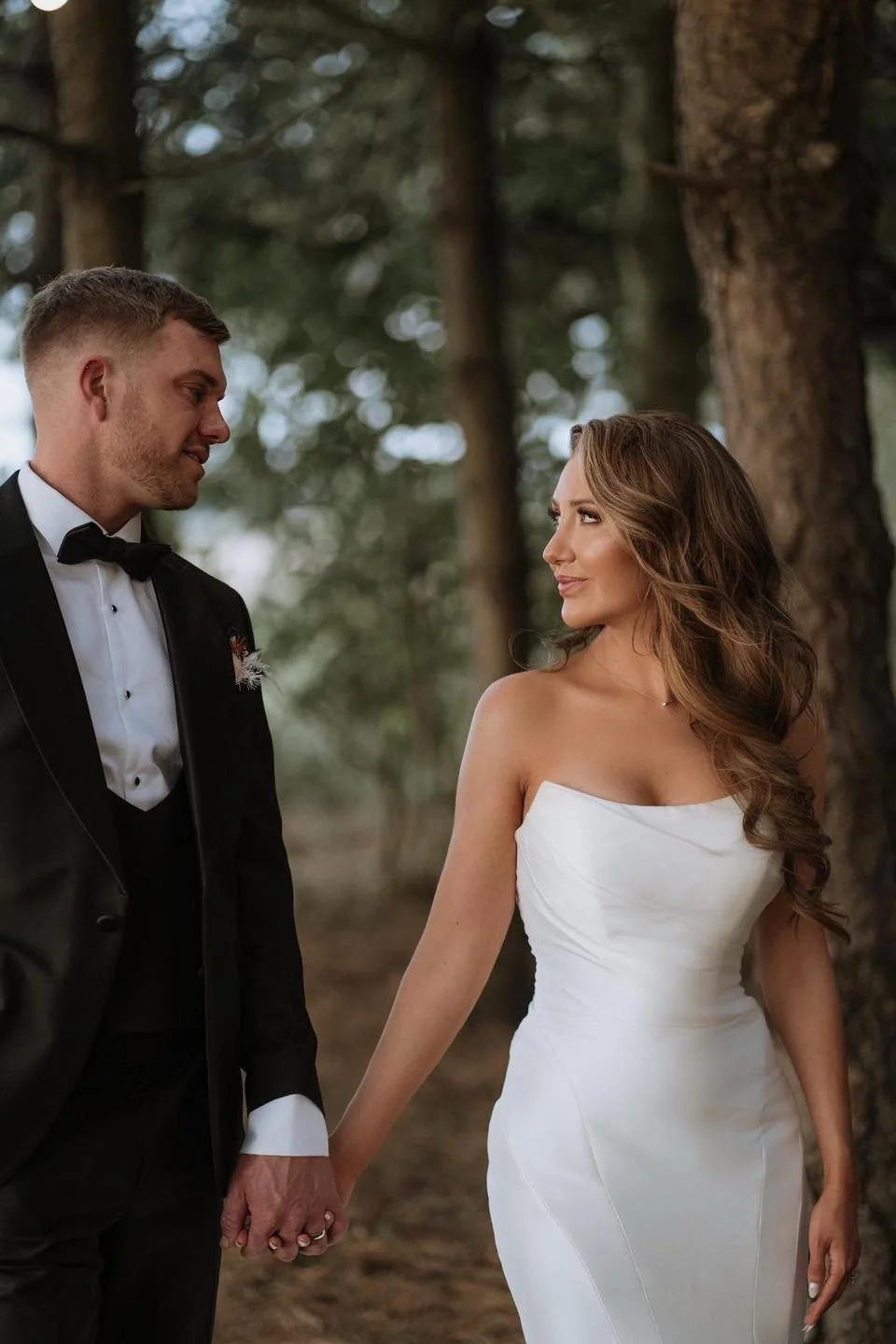 A bride and groom holding hands outside in a wooded area, gazing at each other, with trees and soft natural light in the background.