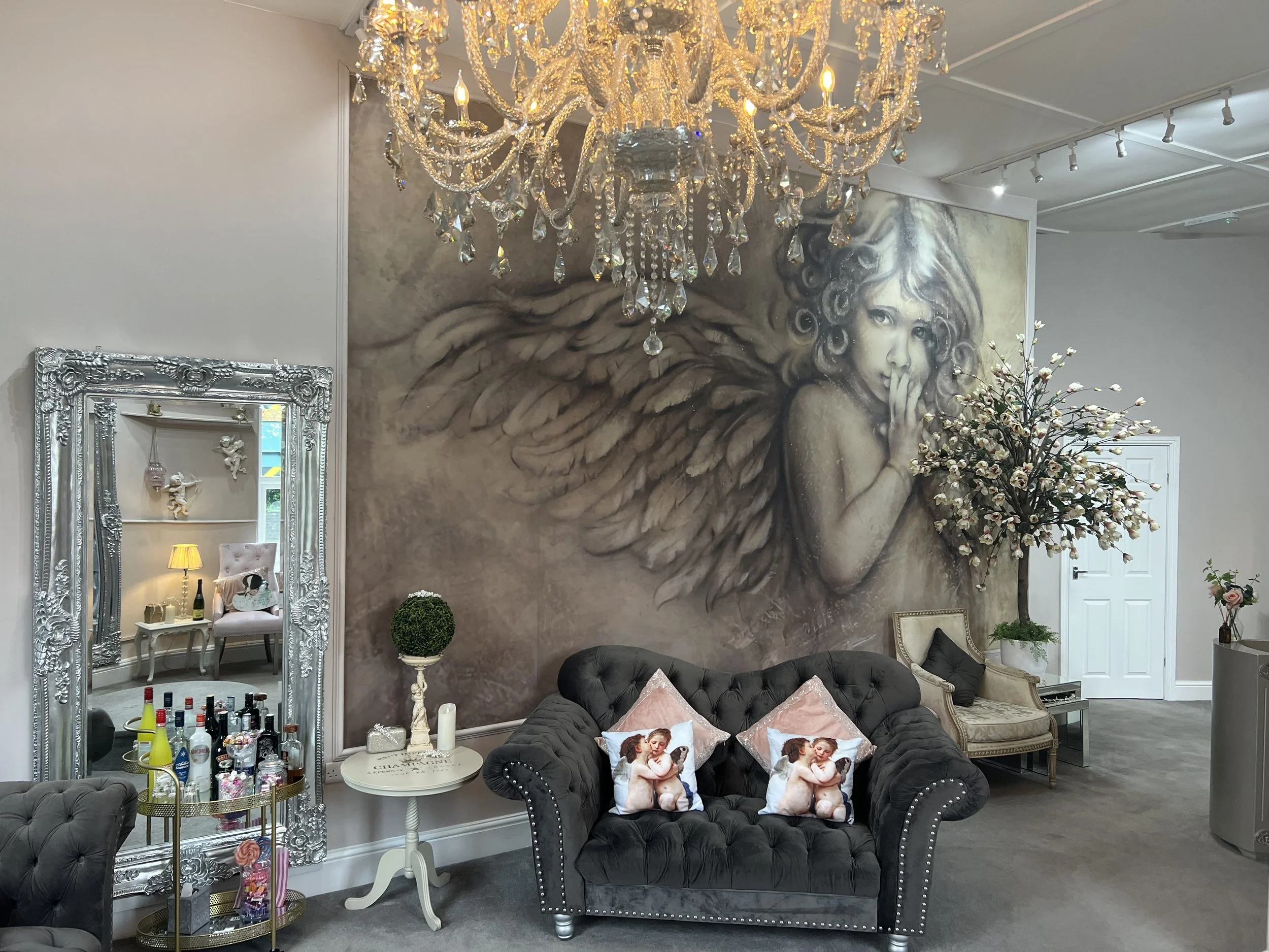 An elegant room with a large chandelier hanging from the ceiling, a detailed silver-framed mirror, a tufted dark sofa with decorative pillows, a small side table, and a wall mural of a winged girl with curly hair. There is a bloom-shaped flower arran