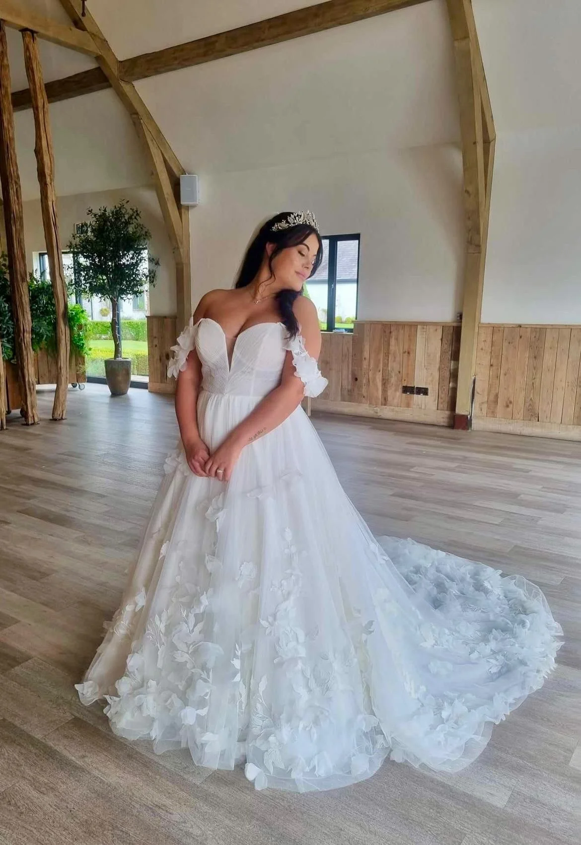 A bride in a white wedding gown with floral details, standing indoors in a rustic-style venue with wooden beams and a large window.
