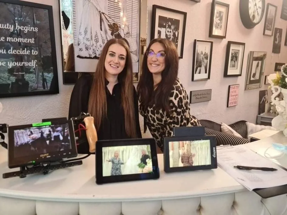 Two women standing behind a reception desk, smiling at the camera, with framed photos and quotes on the wall behind them, and electronic devices on the desk display images.