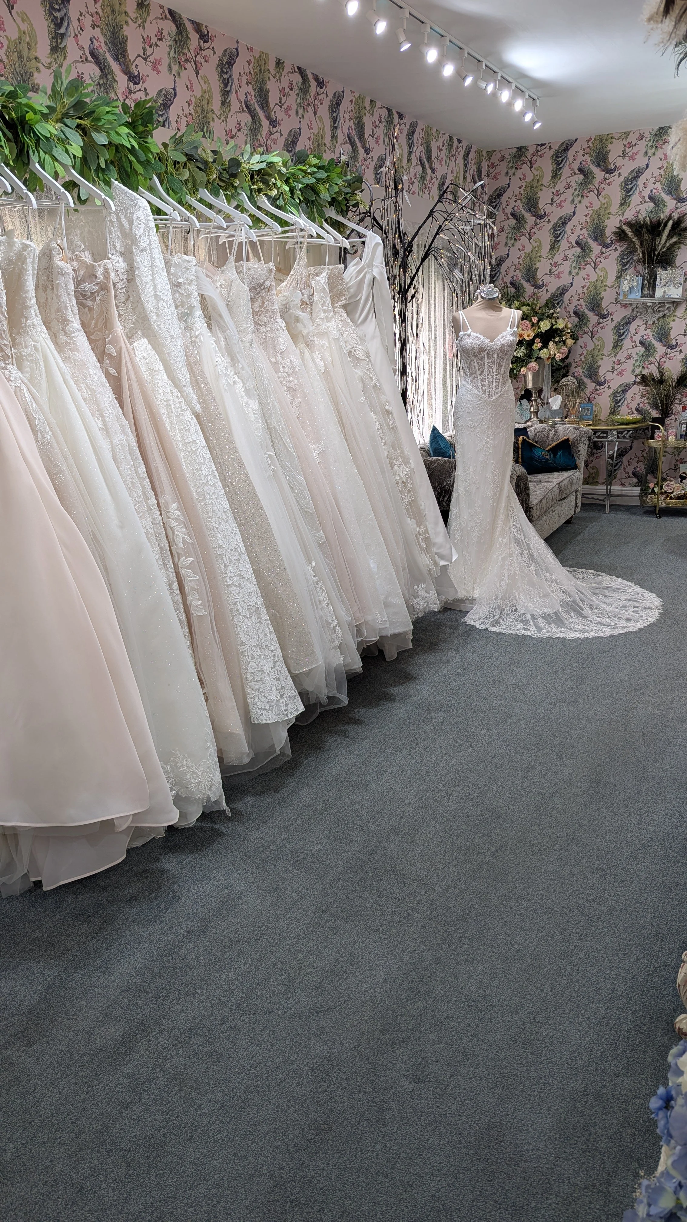Interior of a bridal boutique with a series of white wedding dresses hanging on a rack on the left side. A mannequin wearing a lace wedding gown, displayed near a floral wallpapered wall, is positioned towards the center background. Decorative furnit