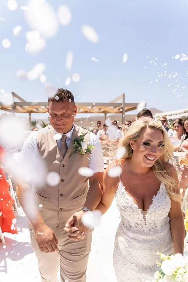 A newlywed couple celebrating outdoors on a sunny day, surrounded by guests, with confetti falling around them.