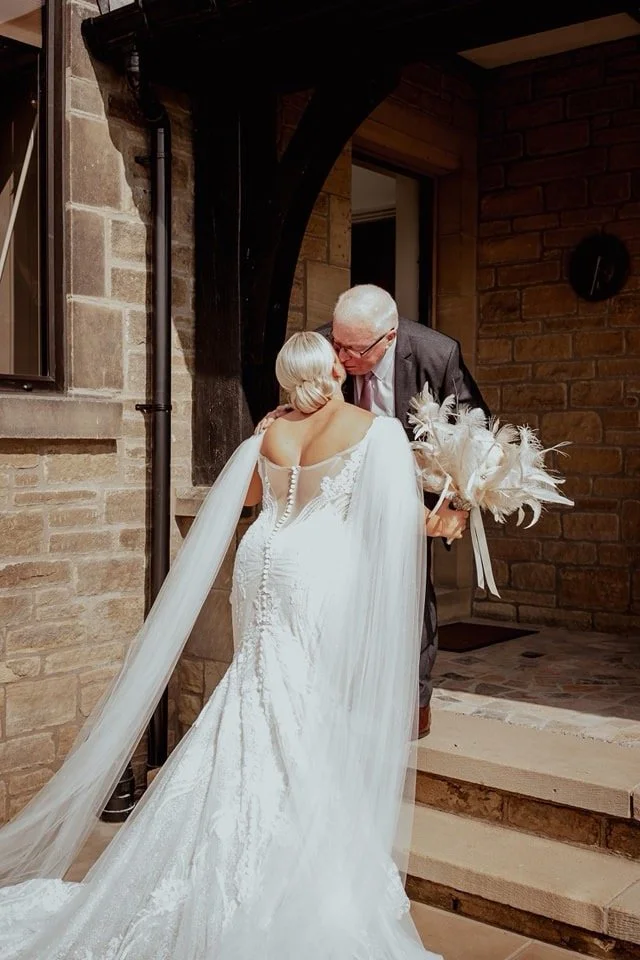 A bride in a white wedding dress with a long train and veil kisses an older man, possibly her father, who is holding a bouquet of white feathers and flowers in front of a brick house.