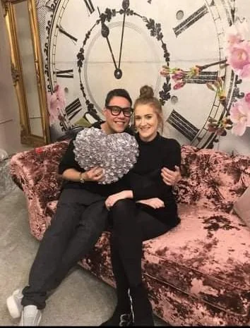 A man and woman sitting on a pink velvet sofa in front of a large decorative clock with floral accents. The man is holding a heart-shaped pillow and both are smiling at the camera.