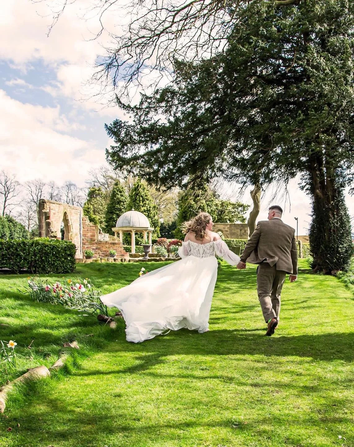 A bride and groom holding hands and running through a lush green garden with trees and a decorative stone and brick gazebo in the background.