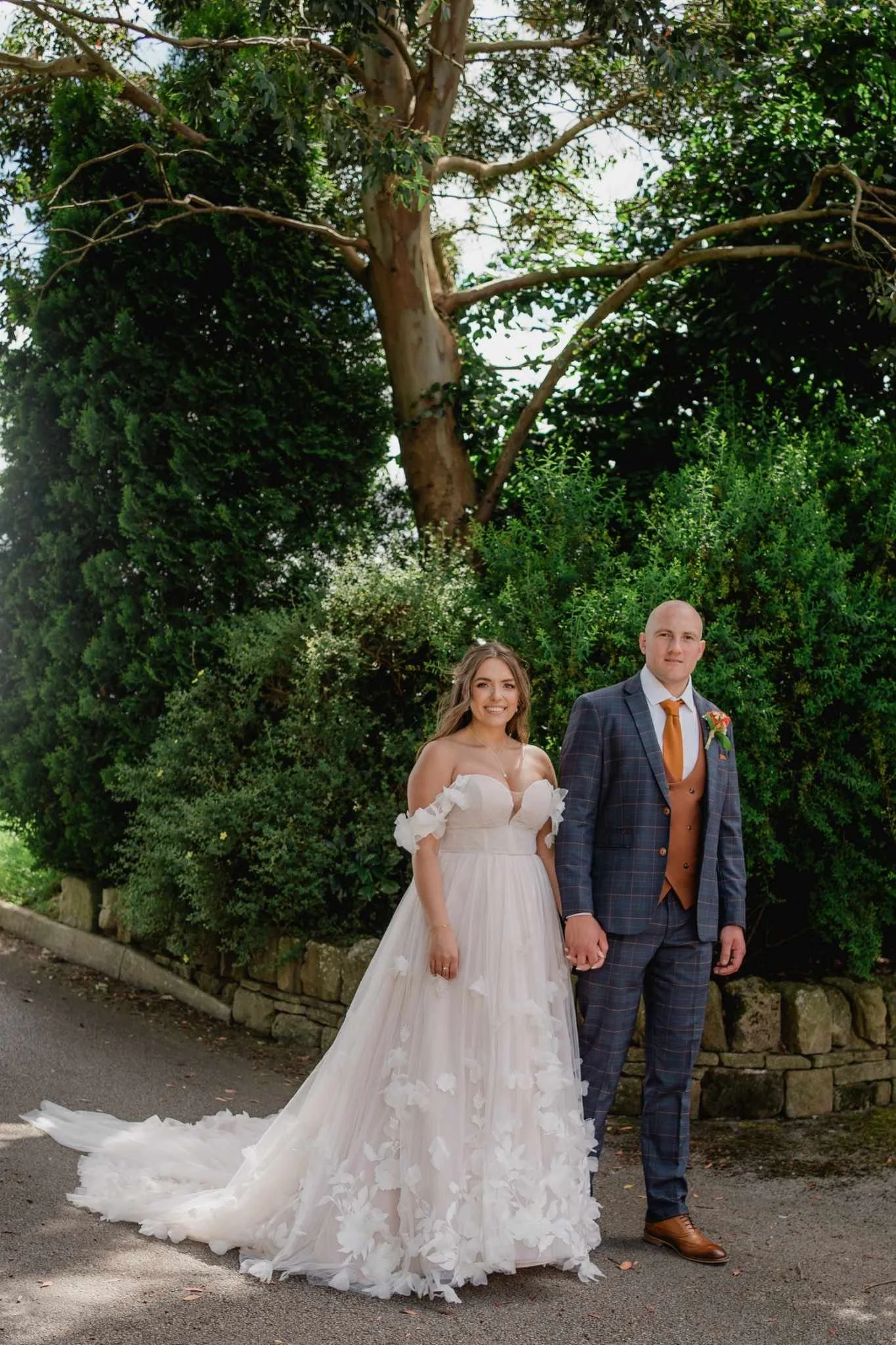 A bride and groom holding hands, standing outdoors on a paved path with trees and bushes in the background. The bride is in a white wedding gown with floral appliqués, and the groom is in a plaid suit with a brown vest and tie.
