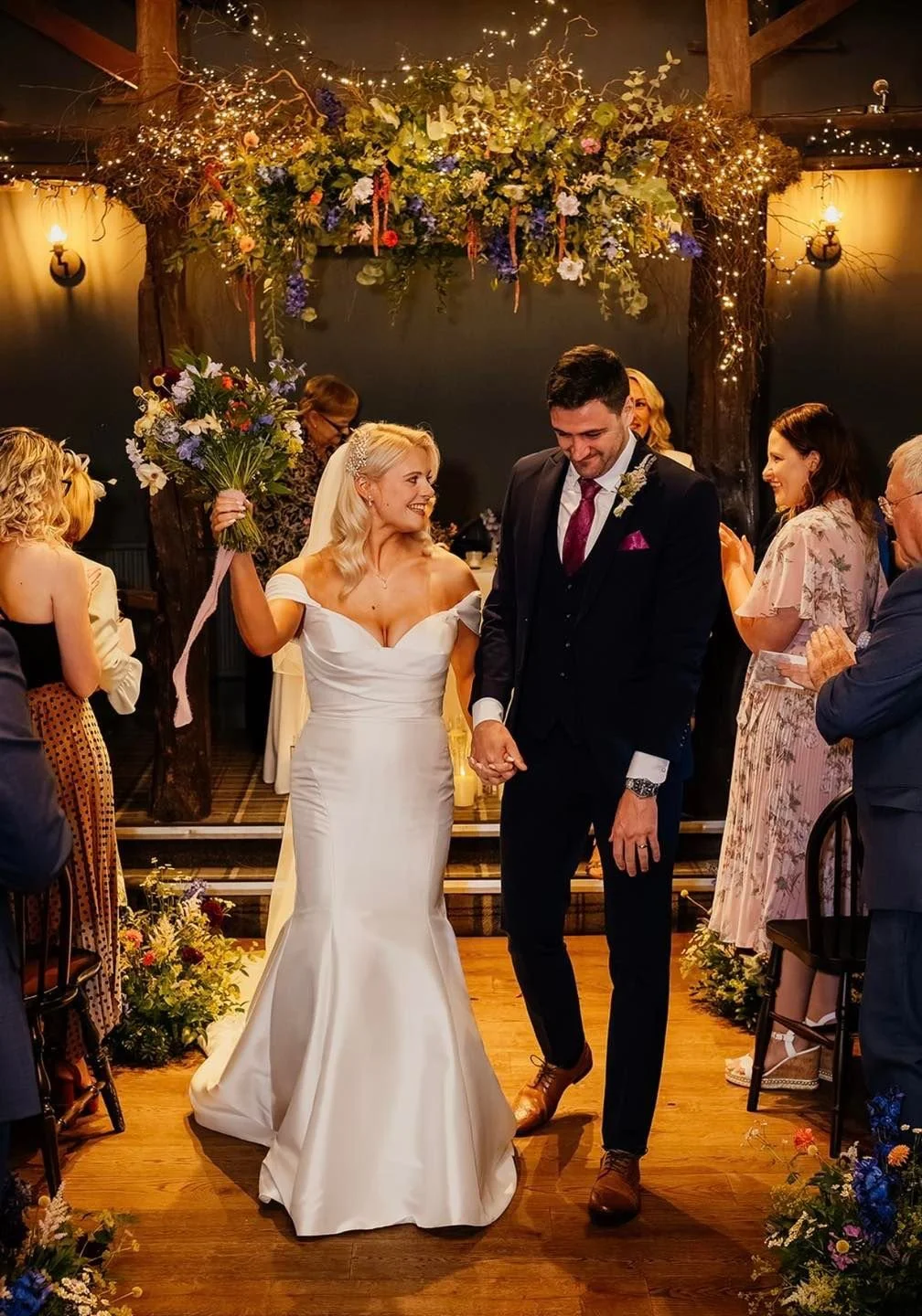 A newlywed couple holds hands walking down the aisle during their wedding ceremony. The bride wears a white off-shoulder wedding gown and holds a bouquet of flowers, smiling at the groom. The groom wears a black suit with a pink tie and boutonniere,
