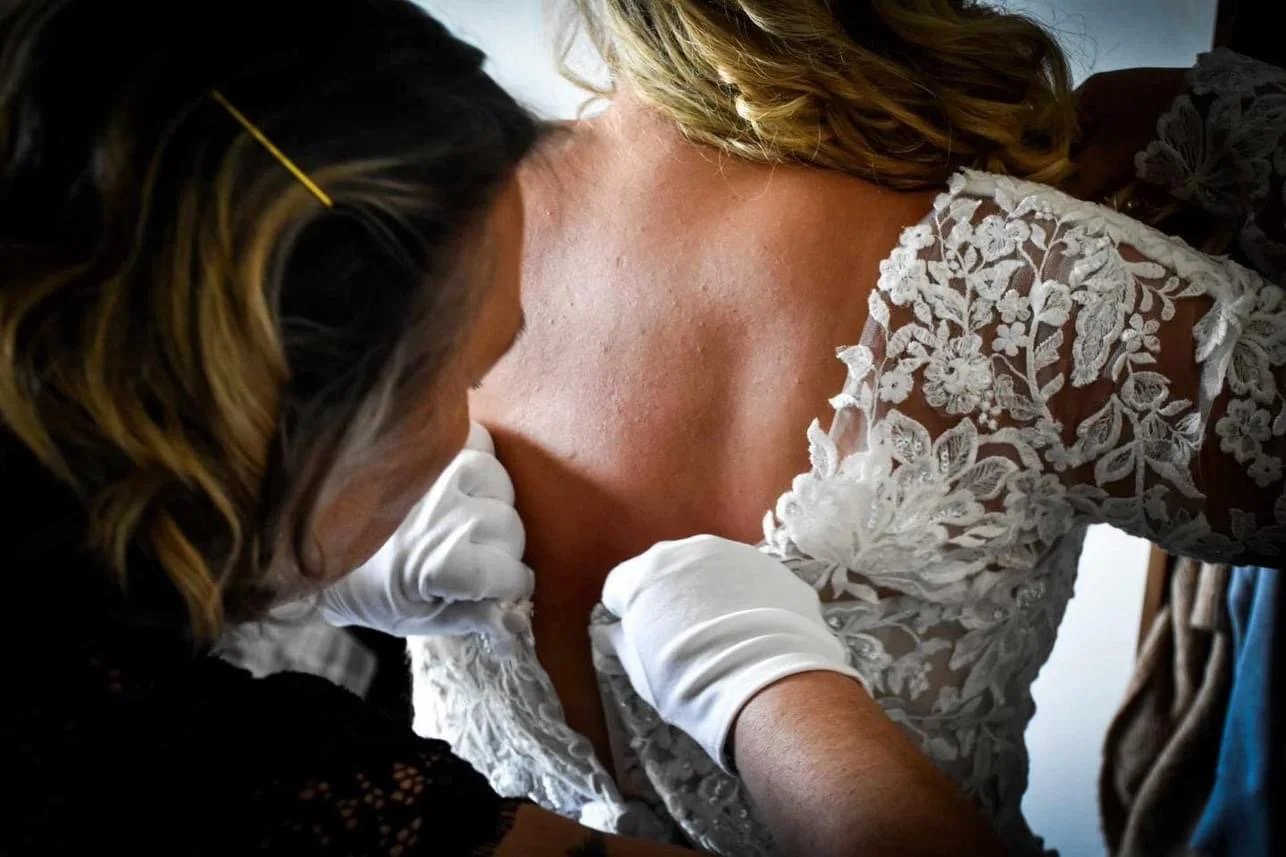 A bride with fair skin and blonde hair in a lace wedding dress is having her dress adjusted by a woman with dark hair, wearing white gloves.