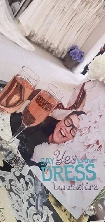 A woman with glasses and a big smile, lying on a table, is surrounded by two glasses of rosé wine, a large floral arrangement, and a sign that reads "Say Yes to the Dress Lancashire," with a background of curtains and flowers.
