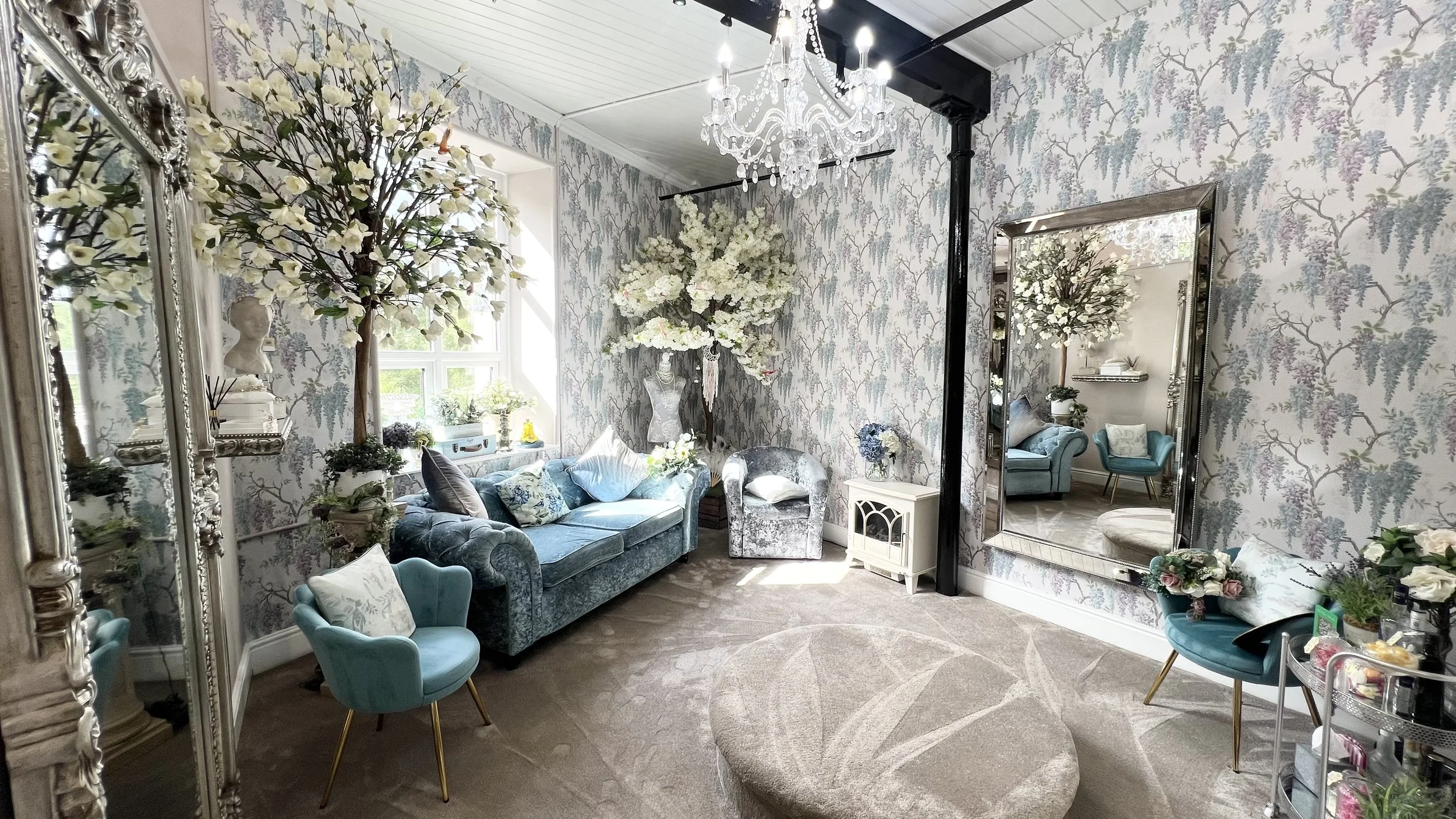 A cozy, elegant living room with vintage-style furniture, floral wallpaper, artificial trees with white blossoms, and a large mirror reflecting part of the room. There's a chandelier hanging from the ceiling and a round ottoman on a patterned beige r